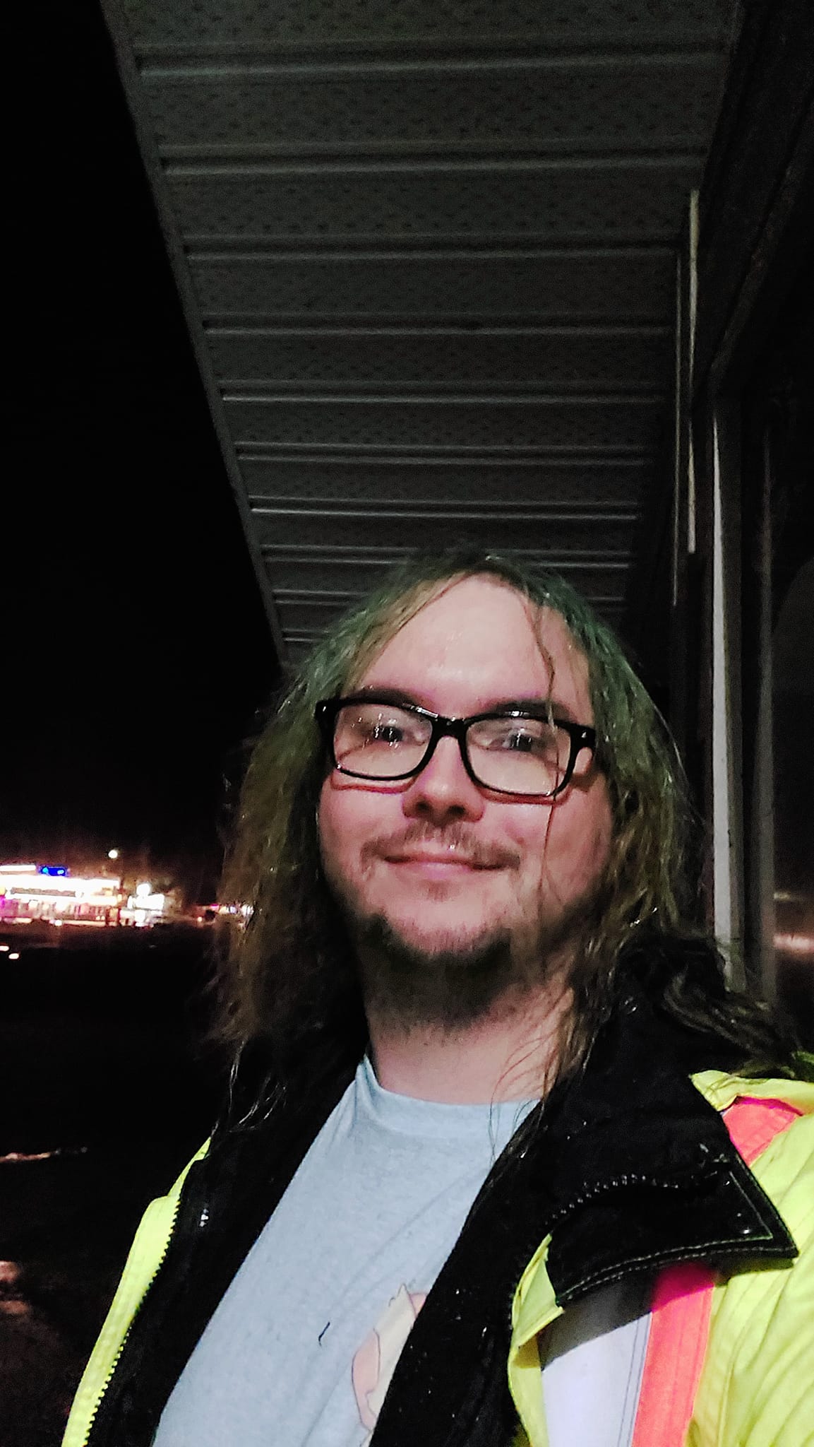 A man with long green hair and no beard wears a high visibility raincoat on a dark night, his hair is wet